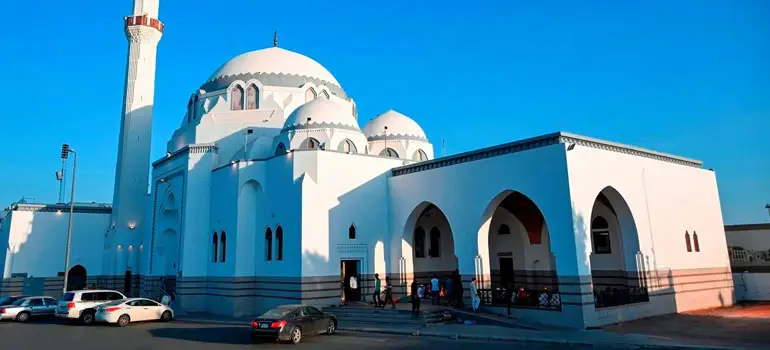 A large white mosque with domes and minaret under a clear blue sky, with parked cars and people outside in Saudi Arabia.