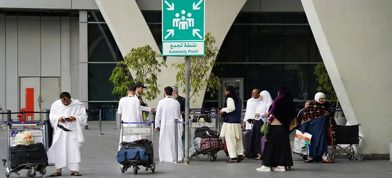 Travelers in traditional Saudi attire with luggage carts gather at an assembly point outside a modern airport terminal in Saudi Arabia.