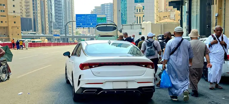 White sedan parked on a busy airport road in Saudi Arabia, surrounded by travelers in traditional clothing carrying luggage near terminal buildings.