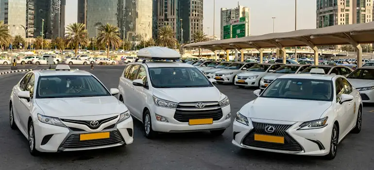 Three white licensed taxis (Toyota Camry, Toyota Innova, and Lexus) parked in a row at a designated taxi stand in a modern Saudi Arabian airport or city with skyscrapers and palm trees in the background.