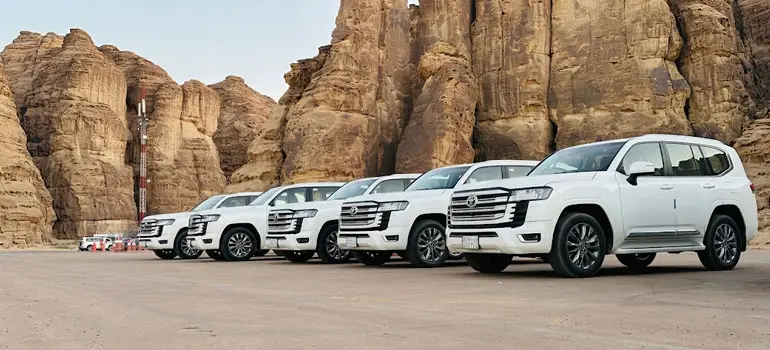 Fleet of white Toyota Land Cruiser SUVs parked in a row against massive sandstone mountain cliffs in Al Ula.