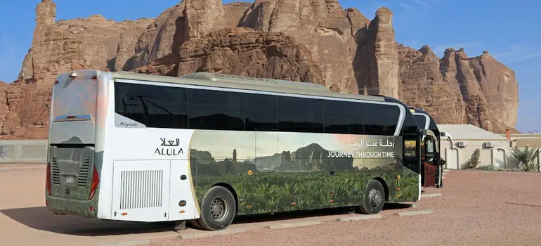 Official Al Ula tourist coach bus labeled parked in front of dramatic desert rock formations.