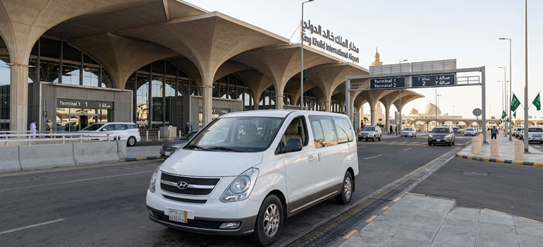 White private transfer van at King Khalid International Airport terminal in Riyadh for airport transport service.