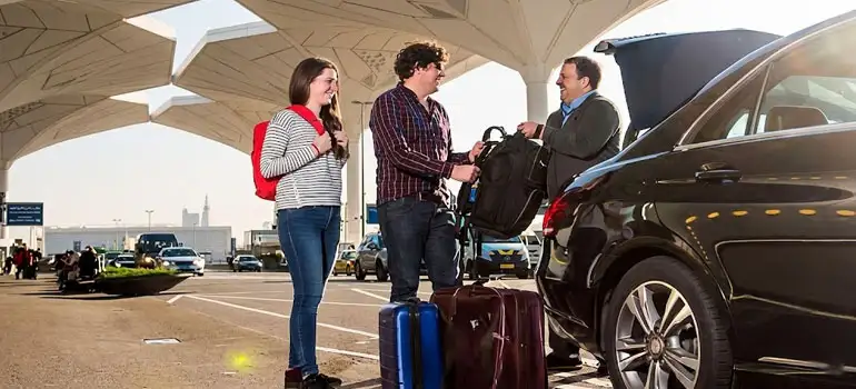 Three travelers—a smiling woman with a red backpack and a man with curly hair—stand with luggage at Riyadh International Airport as a professional driver in a suit loads their bags into the trunk of a black sedan during a sunny pickup.