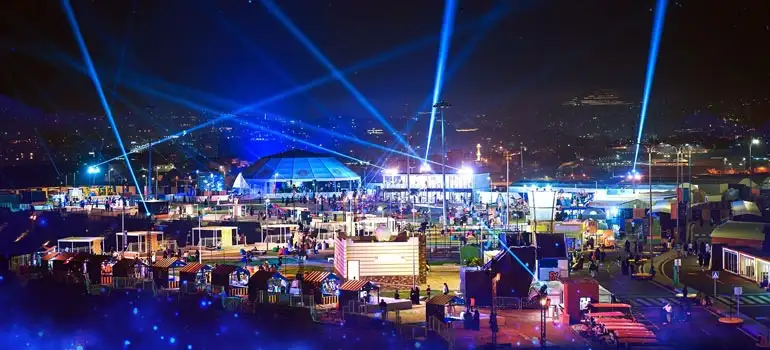 A vibrant nighttime festival in Saudi Arabia with crowds enjoying live entertainment, glowing stages, and dramatic blue laser lights beaming across the dark sky over a brightly lit cityscape.