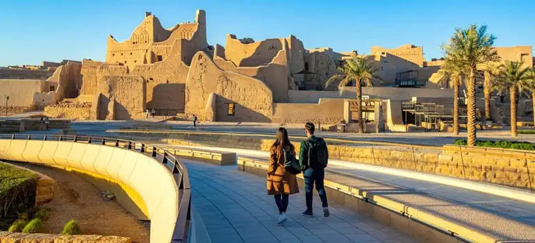 A couple walks along a modern curved promenade with palm trees towards the historic mud-brick fortress of At-Turaif in Diriyah, Riyadh, under a bright sunny sky.